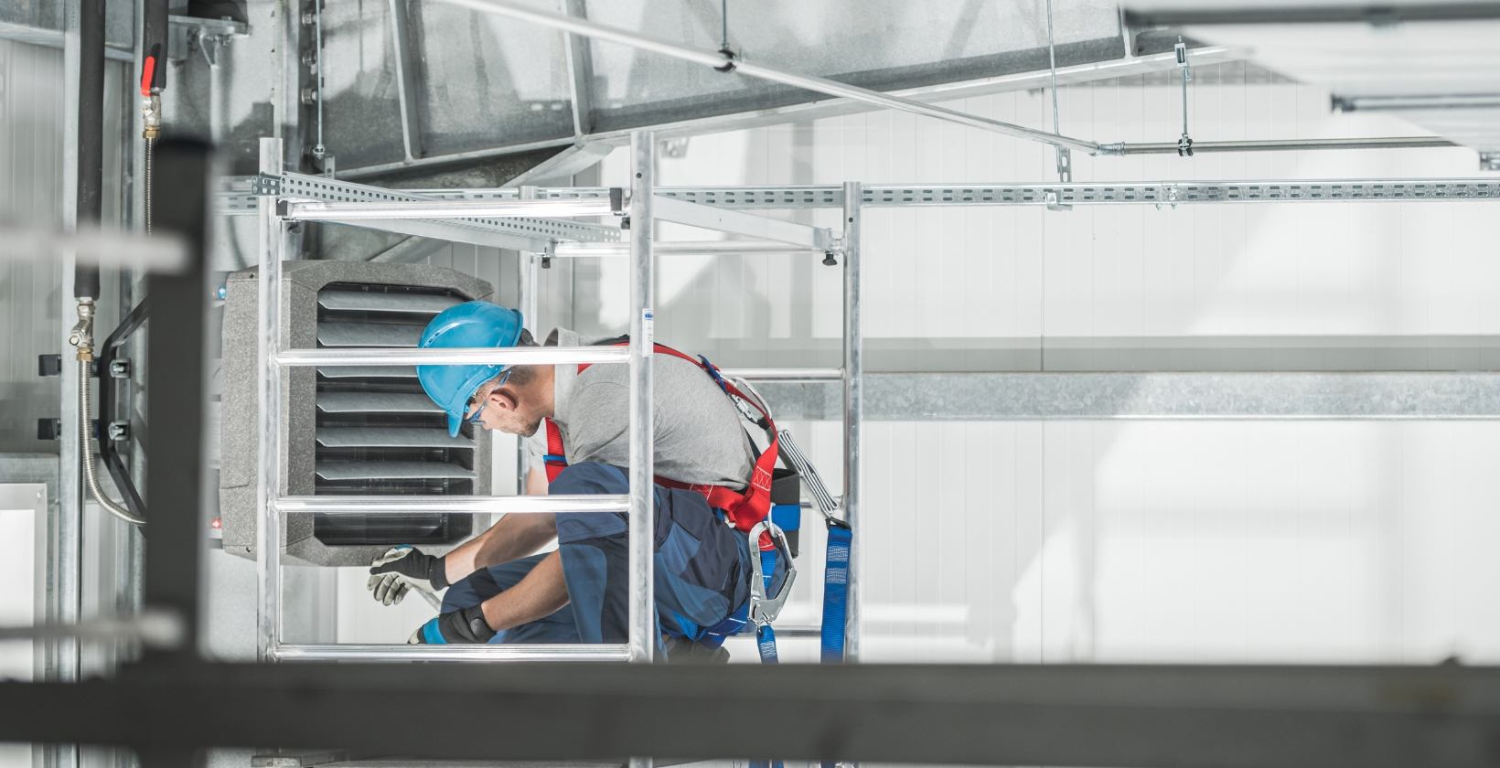 Emergency Gas Repairs | Commercial Gas Pipework Engineer in hard hat and safety harness repairing a wall-mounted warehouse heater from an access tower